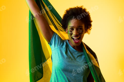 Preview: Image of african american female soccer fan with flag of brazil cheering in yellow lighting