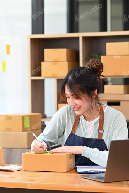 Preview: Pretty young woman writing address on cardboard box while preparing parcel boxes.