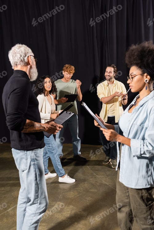 Preview: multiethnic actors holding clipboard with scenarios near grey haired screenwriter in theater