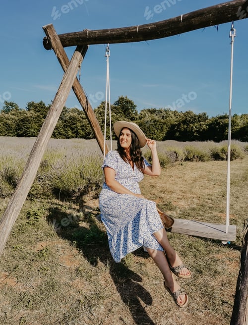 Preview: Vertical shot of a young woman wearing a hat on a wooden swing in the middle of a lavender field
