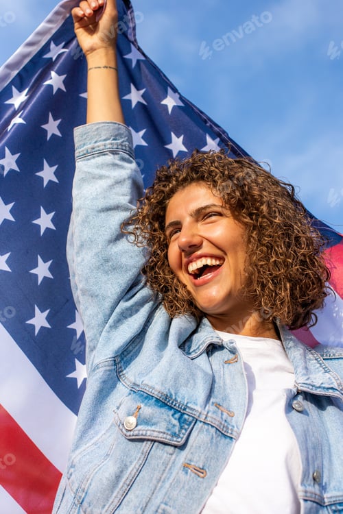 Preview: Happy Beautiful diverse woman holding United States of America flag and standing against blue sky