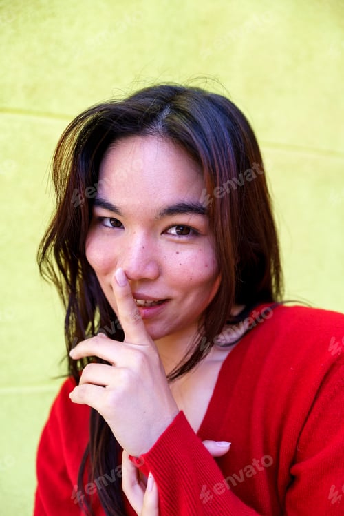 Preview: Young woman wearing red sweater pressing index finger against lips, signaling silence