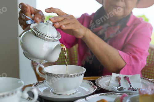 Preview: Close up of an Asian elderly woman in pink blouse pouring hot tea in white porcelain cup