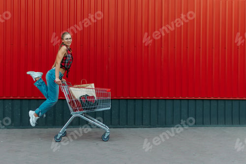 Preview: Happy girl with shopping cart on red wall shop background. Young woman pushing a shopping cart full