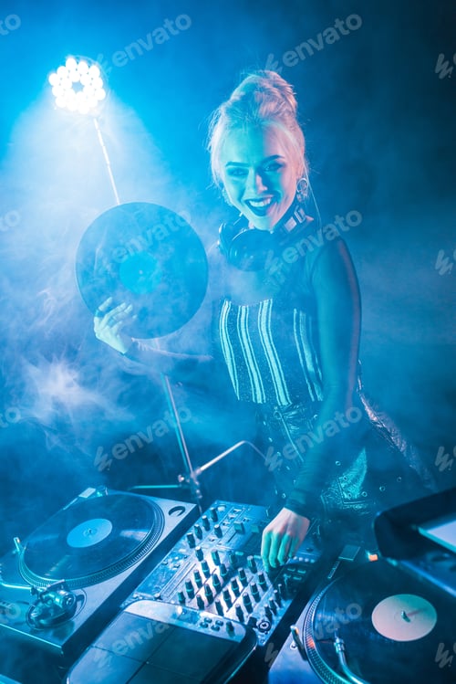 Preview: smiling blonde dj girl touching dj equipment and holding retro vinyl record in nightclub with smoke