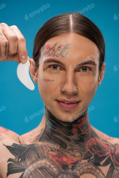 Preview: tattooed young man holding eye patch and smiling while looking at camera isolated on blue
