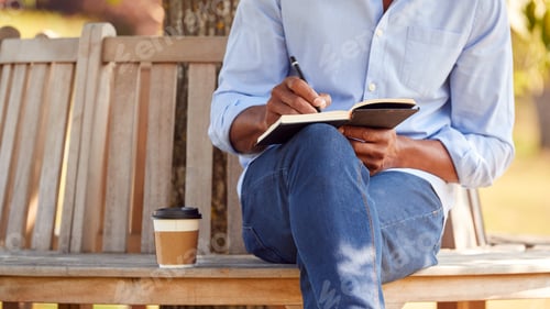 Preview: Close Up Of Man With Takeaway Coffee Sitting On Park Bench Under Tree Writing In Notebook
