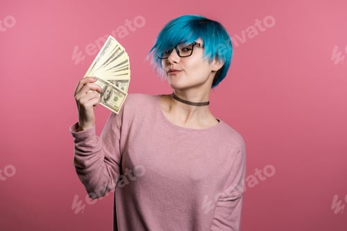 Preview: Satisfied cool excited girl with blue hair showing money - U.S. currency dollars banknotes on pink