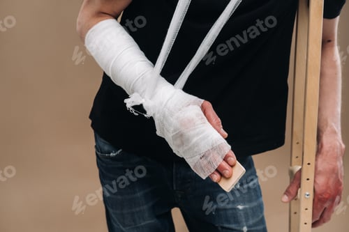 Preview: A battered man with a bandaged head and a cast on his arm stands on crutches on a gray background
