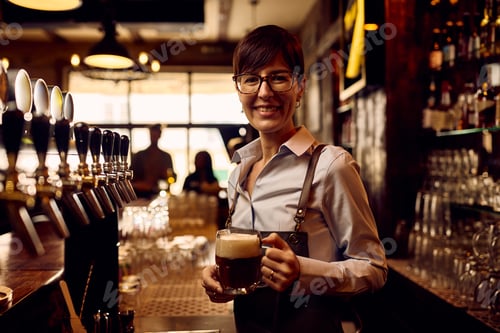 Preview: Happy waitress pouring beer draft beer at bar counter and looking at camera.