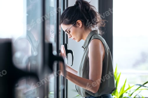 Preview: Worried businesswoman standing while leaning her head against the window at the office.