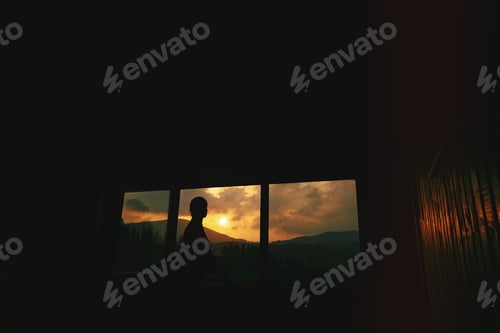 Preview: Silhouette of woman in wooden cottage window with view on sunset in mountains
