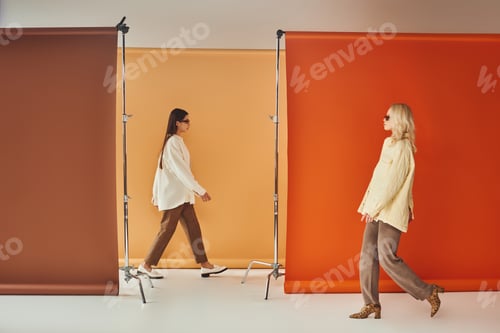 Preview: interracial women in autumn outerwear and sunglasses walking near colorful backdrop, fall fashion