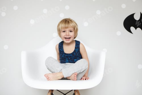 Preview: Portrait of playful toddler with fair hair wearing pajamas, sitting on chair cross-legged, looking a