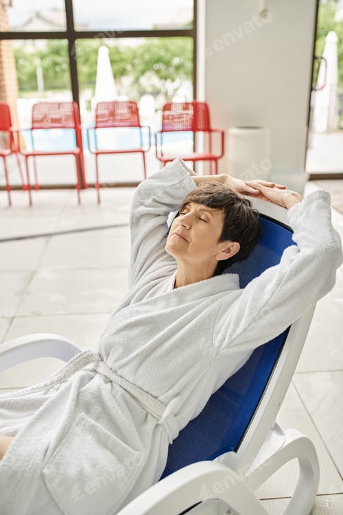 Preview: middle aged woman with short hair relaxing on lounger, white robe, spa center, closed eyes