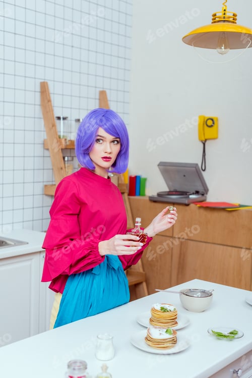 Preview: selective focus of housewife with purple hair and colorful clothes pouring syrup on pancakes in