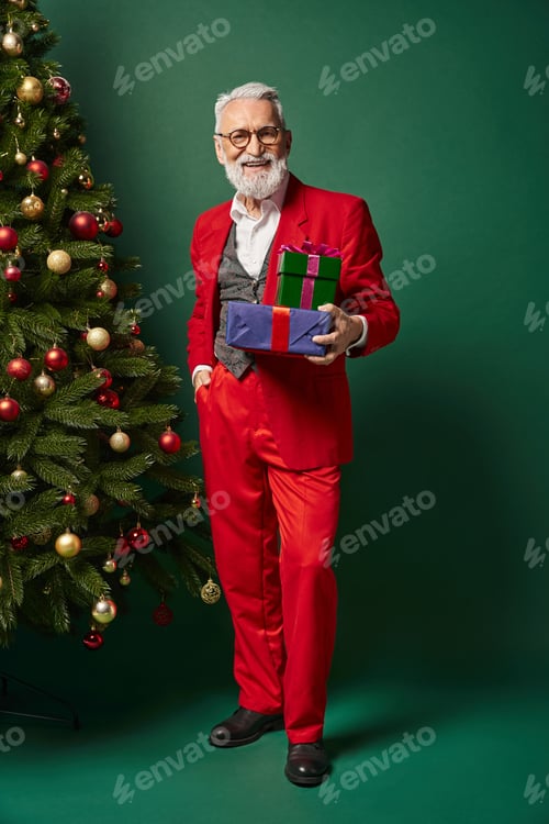 Preview: joyful Santa with glasses posing with presents and one hand in pocket looking at camera, winter