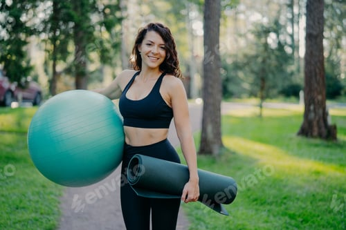 Preview: Sporty European woman in cropped top and leggings, carries rolled up karemat and fitness ball