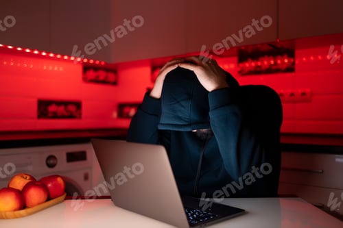 Preview: a hacker in a black hoodie with a hood on his head is sitting at a laptop lowered his head