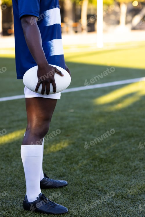 Preview: African American young male athlete holding a rugby ball, standing on field outdoors, copy space