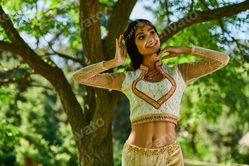 Preview: elegant indian woman in traditional clothes smiling and looking away in green park, summer leisure