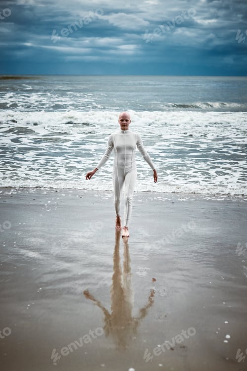 Preview: Hairless girl with alopecia in white futuristic suit comes out of cold restless sea on sandy beach