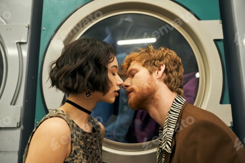 Preview: young and stylish multiethnic couple kissing near washing machine in coin laundry