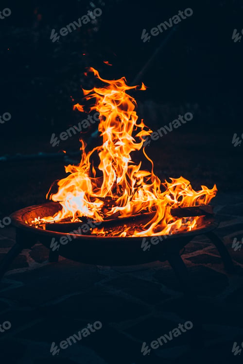 Preview: Vertical shot of a bonfire burning in a metallic bowl on a sandy beach at night