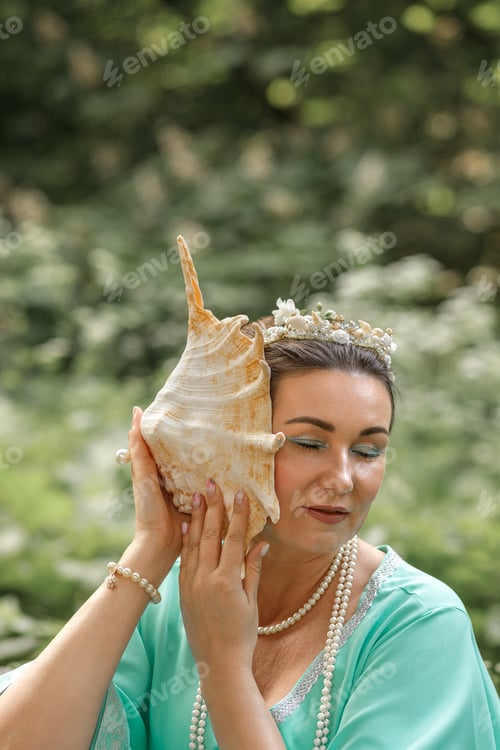 Preview: Beautiful brunette girl in a turquoise long dress in nature by the lake. Siren put a shell