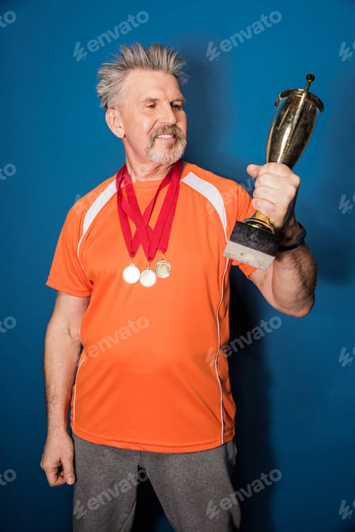 Preview: Smiling mature sportsman with medals holding trophy cup isolated on blue