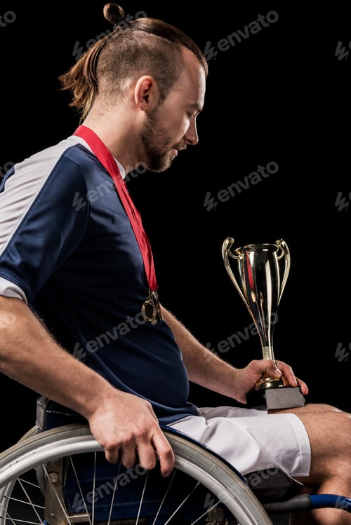 Preview: paralympic in wheelchair with gold medals on neck looking at champion goblet isolated on black