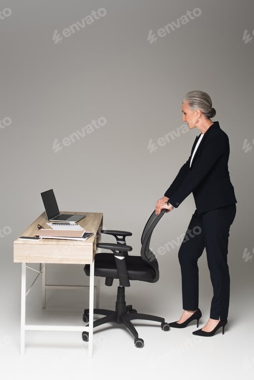 Preview: Side view of mature businesswoman standing near office chair and working table on grey background