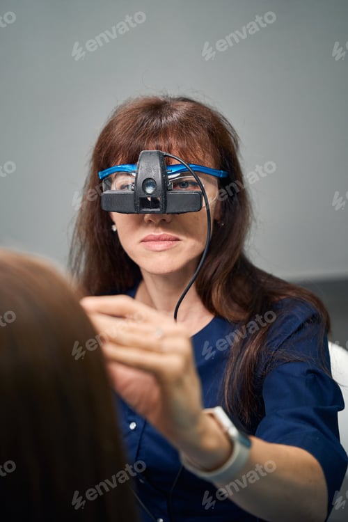 Preview: Female doctor using headgear device, focused on patients eyes