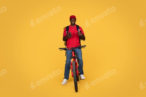Preview: Smiling millennial african american guy courier in uniform with backpack on bike, point finger at