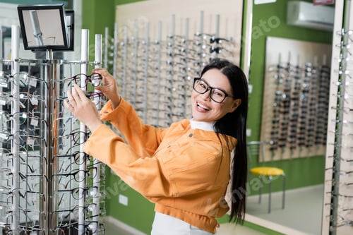 Preview: joyful asian woman smiling at camera while choosing spectacles in optics store