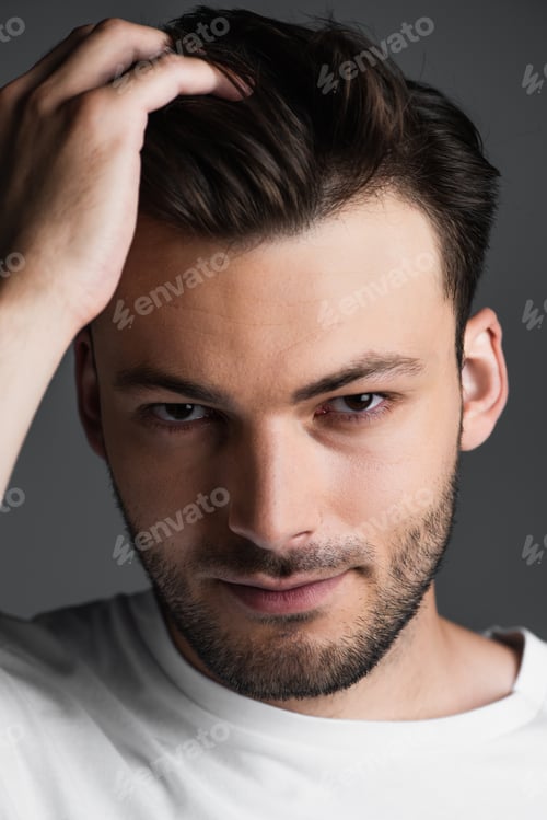 Preview: Portrait of young man touching hair and looking at camera isolated on grey
