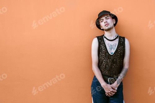 Preview: Tattooed person from the lgbt community in casual attire posing on an orange wall.