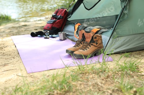 Preview: Boots and camping equipment near tent on riverbank. Space for text
