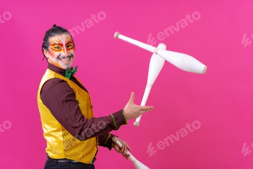 Preview: Clown with white facial makeup on a pink background, smiling juggling clubs and looking at camera