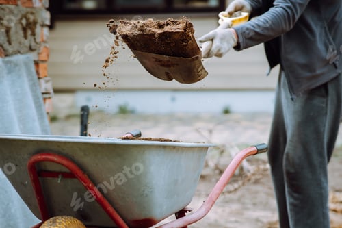 Preview: Human in tracksuit and gloves holding spade and throw earth into wheelbarrow. Gardener removing land