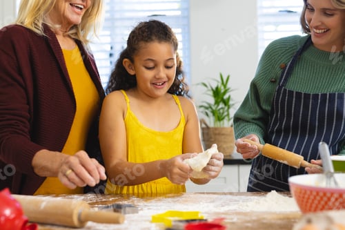 Preview: Happy multiracial grandmother and mother looking at girl kneading dough on table in kitchen