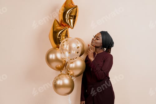 Preview: Muslim black girl holding gold balloons on peach background. African young woman birthday party