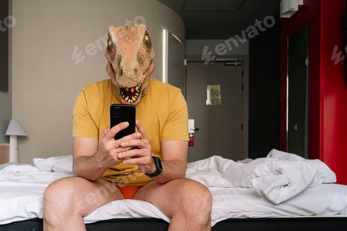 Preview: Man with lizard mask using the smartphone in a hotel bed.