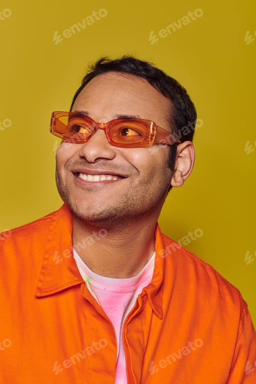 Preview: happy indian man in orange sunglasses looking away while smiling on yellow background, side glance