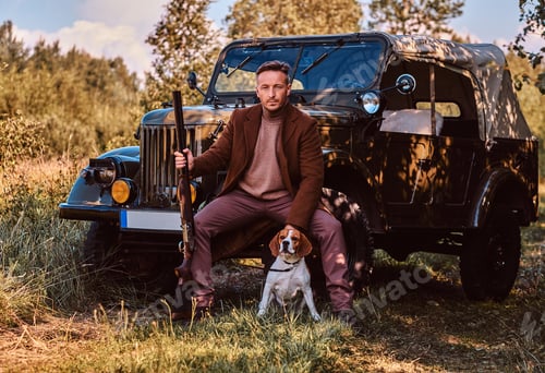 Preview: Hunter in elegant clothes together with his beagle dog next to a retro military car in the forest.