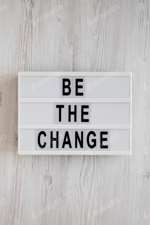 Preview: 'Be the change' words on a modern board on a white wooden background, top view.