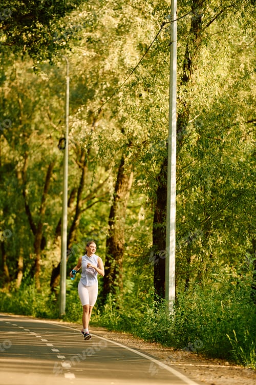 Preview: Sporty young woman jogging through a beautiful green path surrounded by trees in the morning