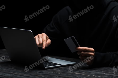 Preview: cropped view of hacker using laptop while holding credit card isolated on black