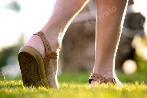 Preview: Close up of woman feet in summer sandals shoes walking on spring lawn covered with fresh green grass