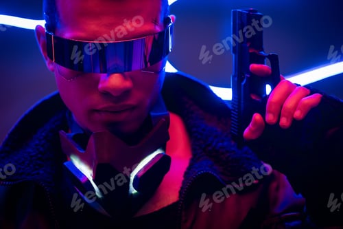 Preview: handsome mixed race cyberpunk player in futuristic glasses holding gun near blue neon lighting